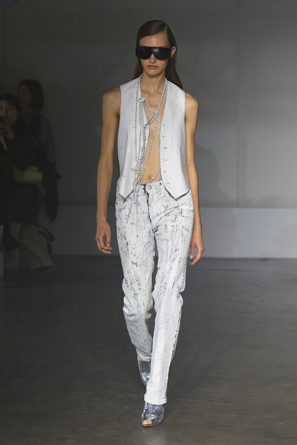 Model walking runway in a sleeveless white vest over distressed pale jeans with pearl chains draped across the torso and oversized black sunglasses, silver ankle boots visible.