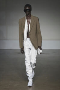 Male fashion model on a runway wearing a tan oversized blazer, white waistcoat, paint-splattered white jeans, white boots, and dark sunglasses, holding a white clutch.