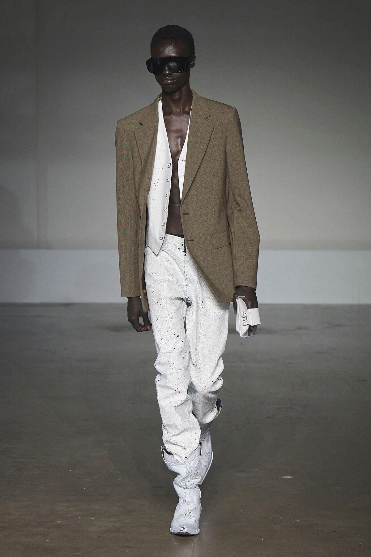 Male fashion model on a runway wearing a tan oversized blazer, white waistcoat, paint-splattered white jeans, white boots, and dark sunglasses, holding a white clutch.