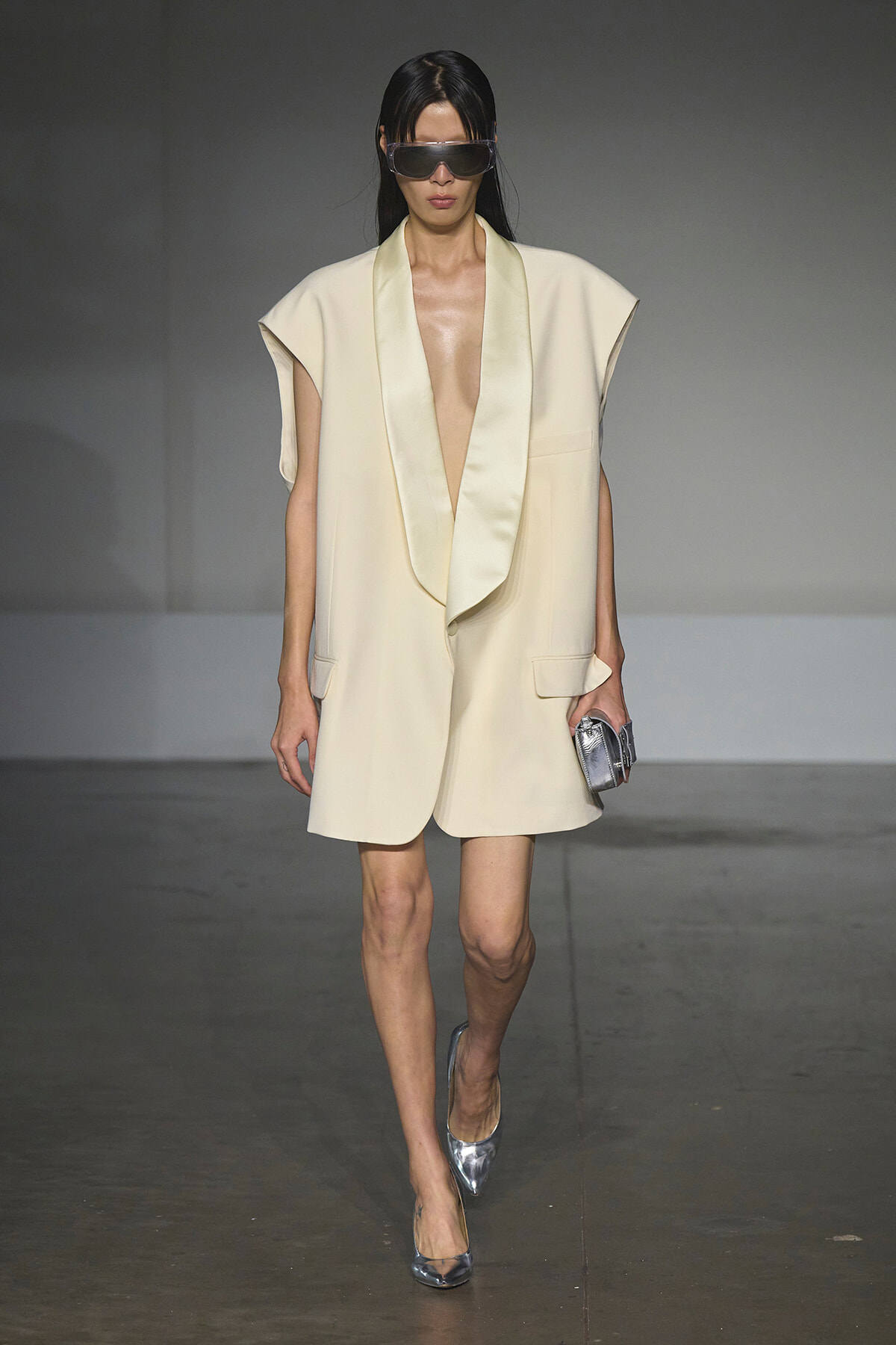 Model on runway wearing a cream sleeveless blazer with a draped shawl lapel, paired with silver heels and a metallic clutch, plus oversized sunglasses.