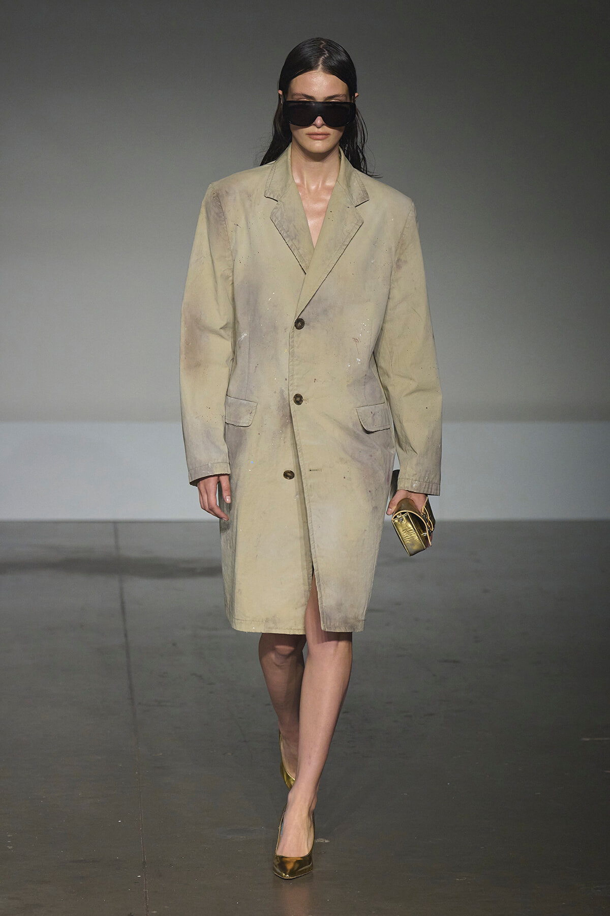 Model walks the runway in a long, oversized beige coat with paint splatters, paired with black sunglasses and gold heels, carrying a small gold clutch.