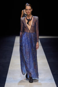 Model on a runway wearing a sheer, glittery blue gown with a deep V neckline and black statement necklace.