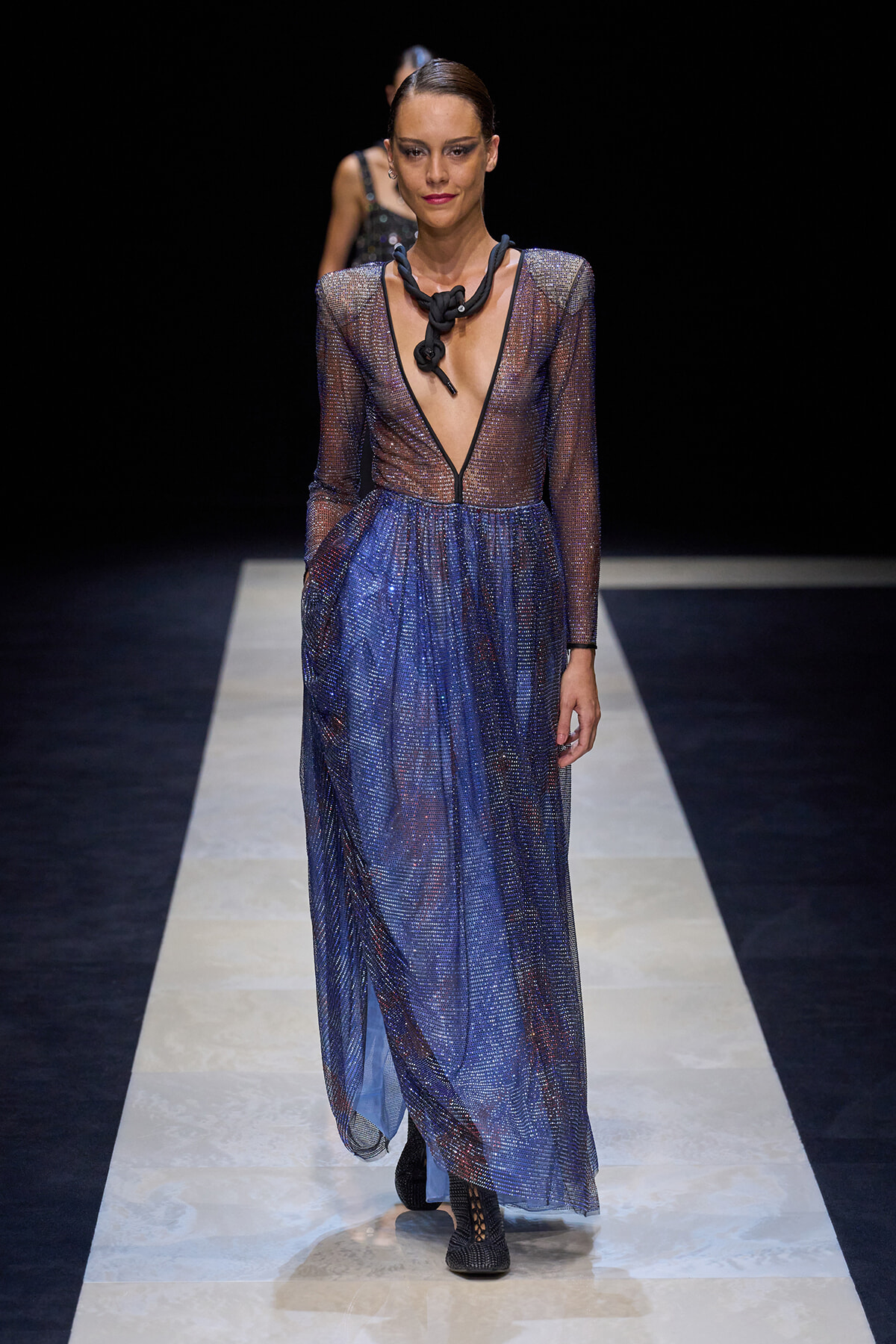 Model on a runway wearing a sheer, glittery blue gown with a deep V neckline and black statement necklace.