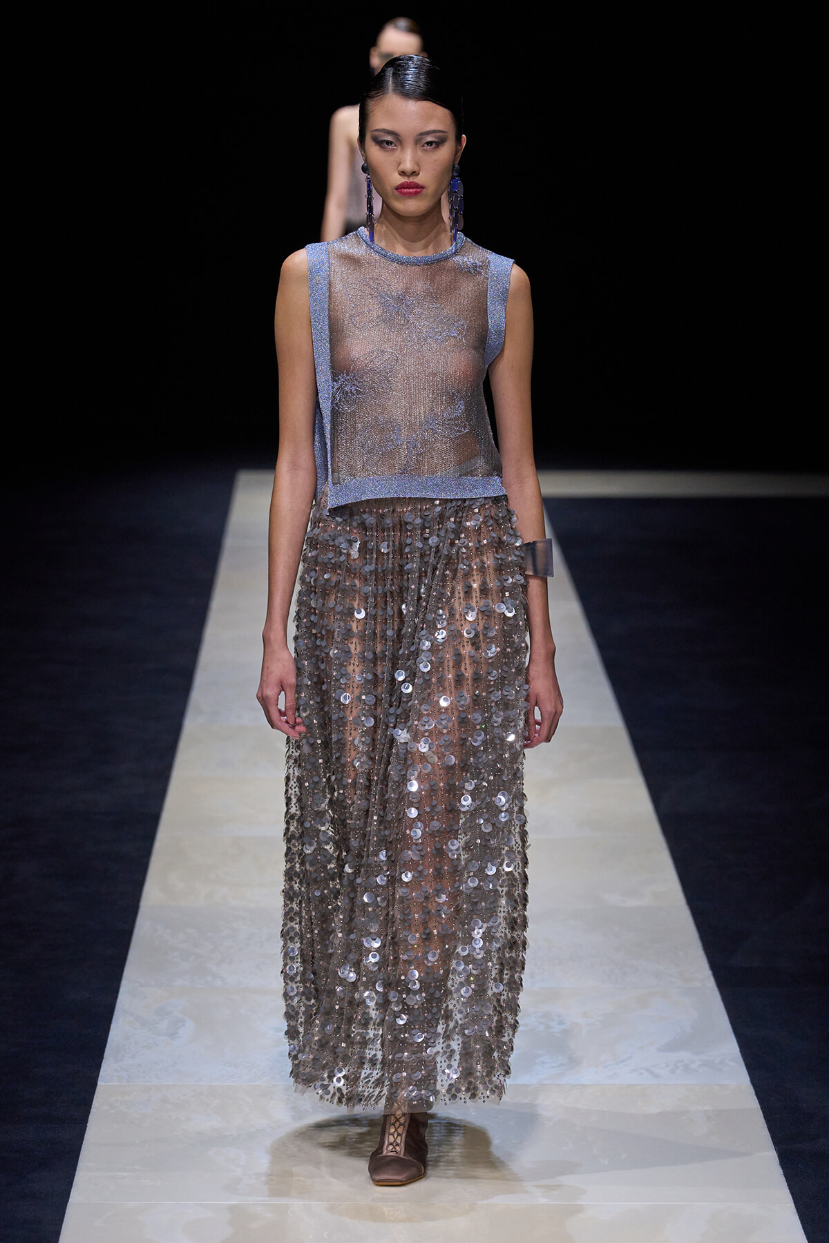 Model on a runway wearing a sheer blue sleeveless top with a metallic-sequin floor-length skirt and brown flat shoes, accessorized with bold blue earrings.