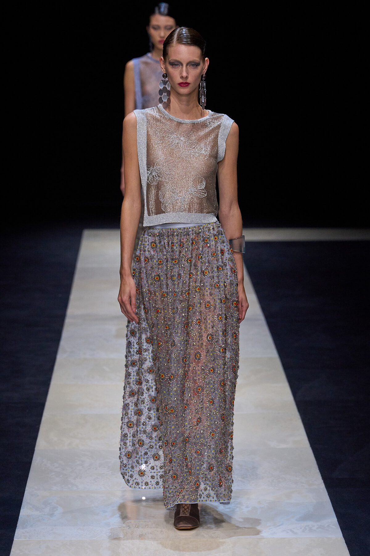 Model on a runway wearing a sleeveless sheer silver top and a floor-length floral-embellished gray skirt with brown ankle boots and bold earrings.