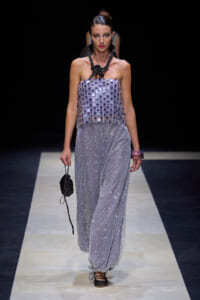 Model walking a fashion show wearing a strapless lavender sequin top and wide, glittery pants; bold black braided necklace and small black pouch in hand.