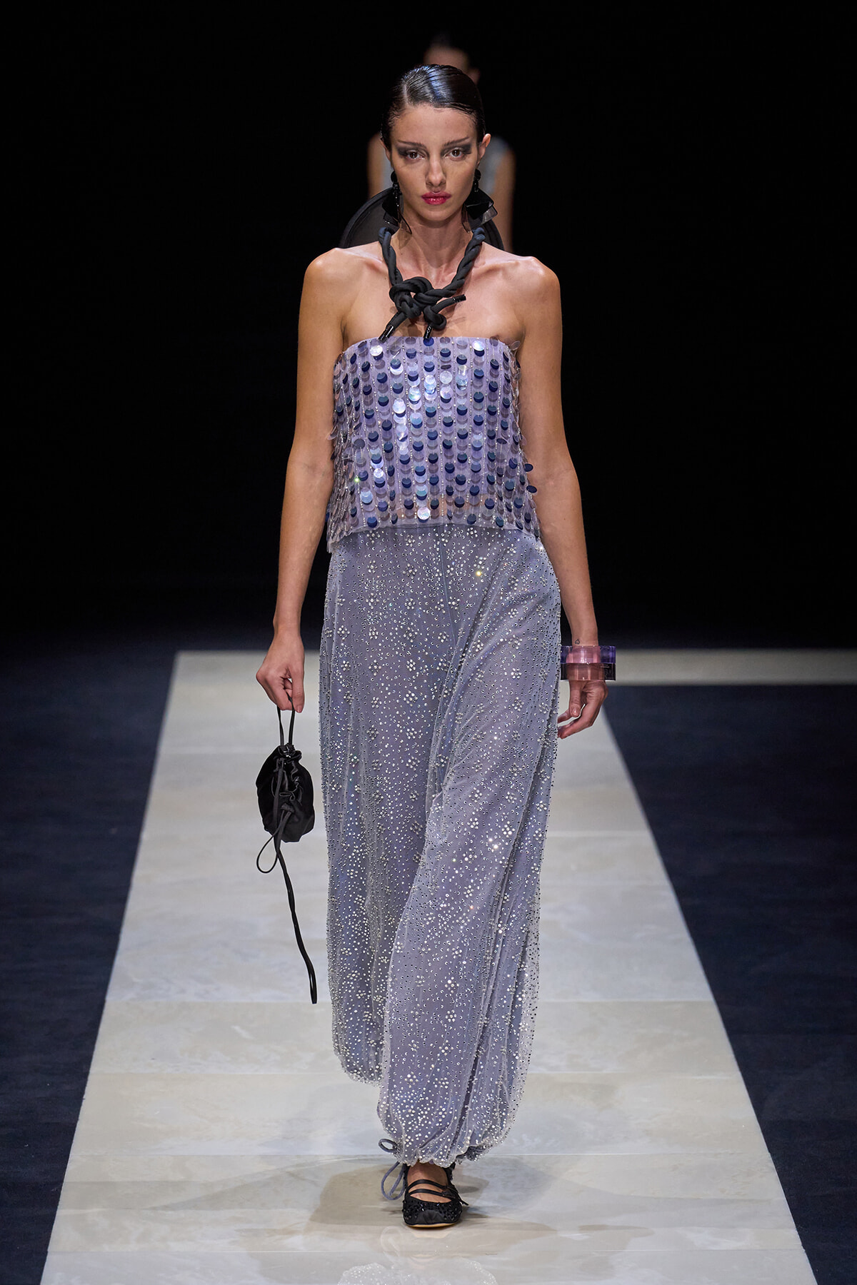 Model walking a fashion show wearing a strapless lavender sequin top and wide, glittery pants; bold black braided necklace and small black pouch in hand.