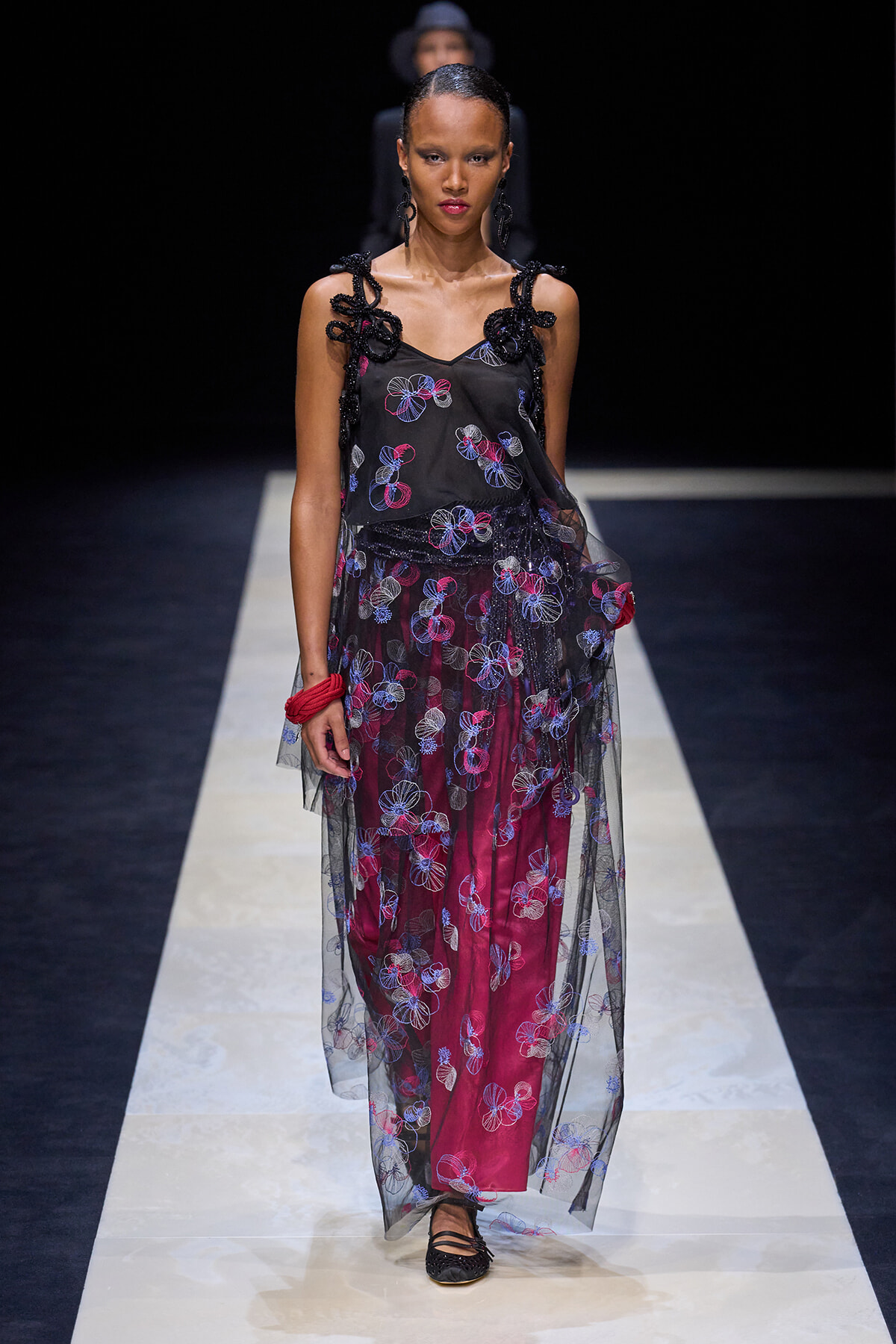 Model walks a fashion runway in a black sheer top with embroidered flowers over magenta pants, covered by a floral tulle overlay.