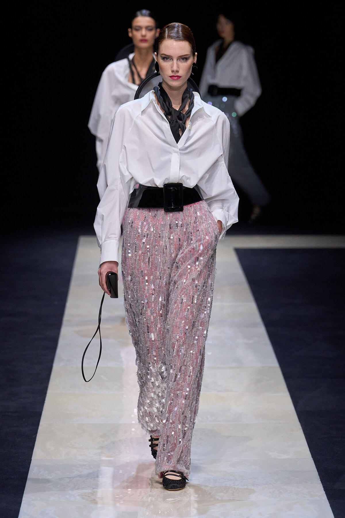 Model walking the runway in a white oversized shirt, pink sequin pants, a wide black belt, and a black neck scarf, carrying a small black clutch.