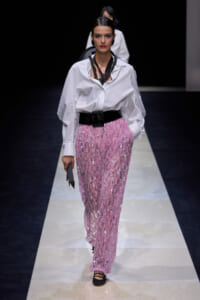 Model walking a runway in a white oversized shirt tucked into a pink sequined skirt with a wide black belt and black accessories.