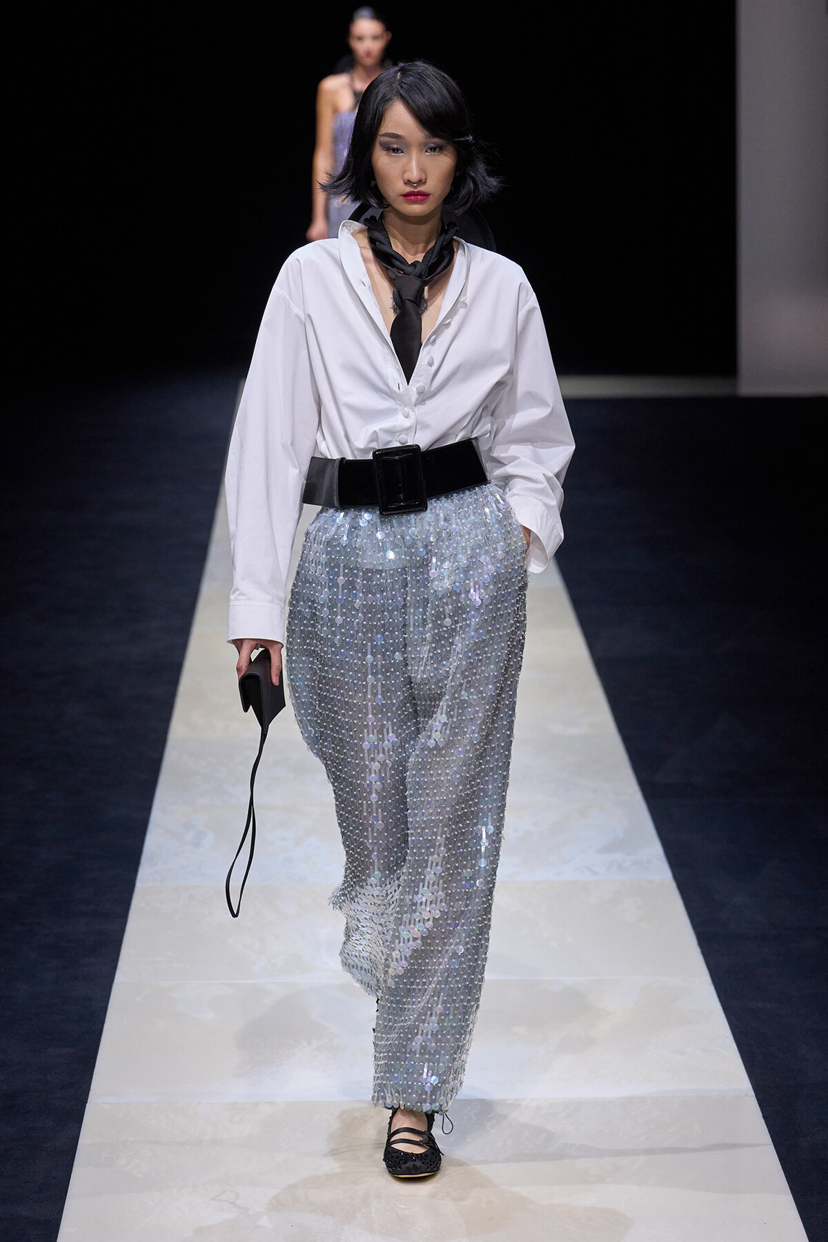 Female model on a runway wearing a white oversized button-down shirt, a wide black belt, and shimmering silver sequined pants, with a black scarf and small clutch.