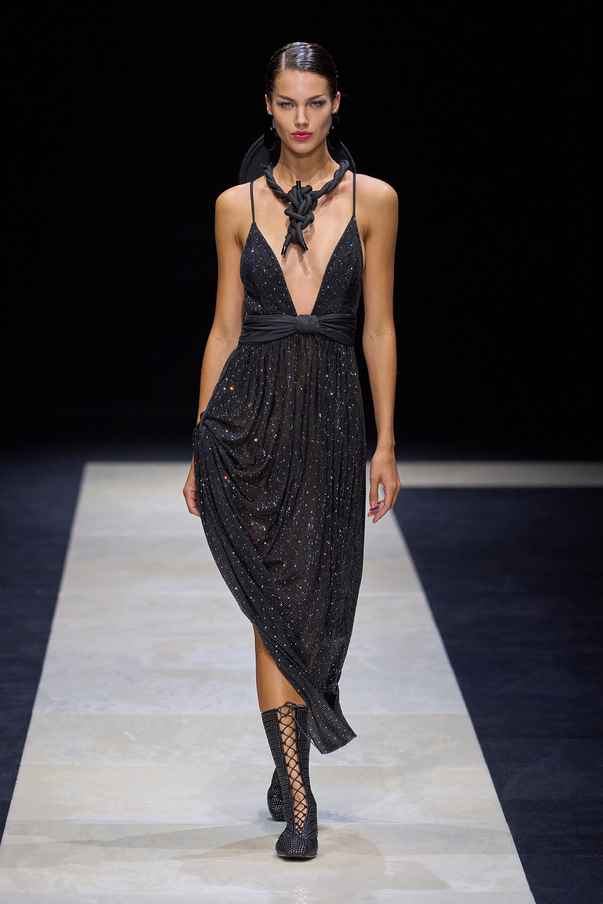 Model walking a runway in a sparkly black evening dress with plunging neckline and wrapped waist, paired with black lace-up boots and bold necklace.