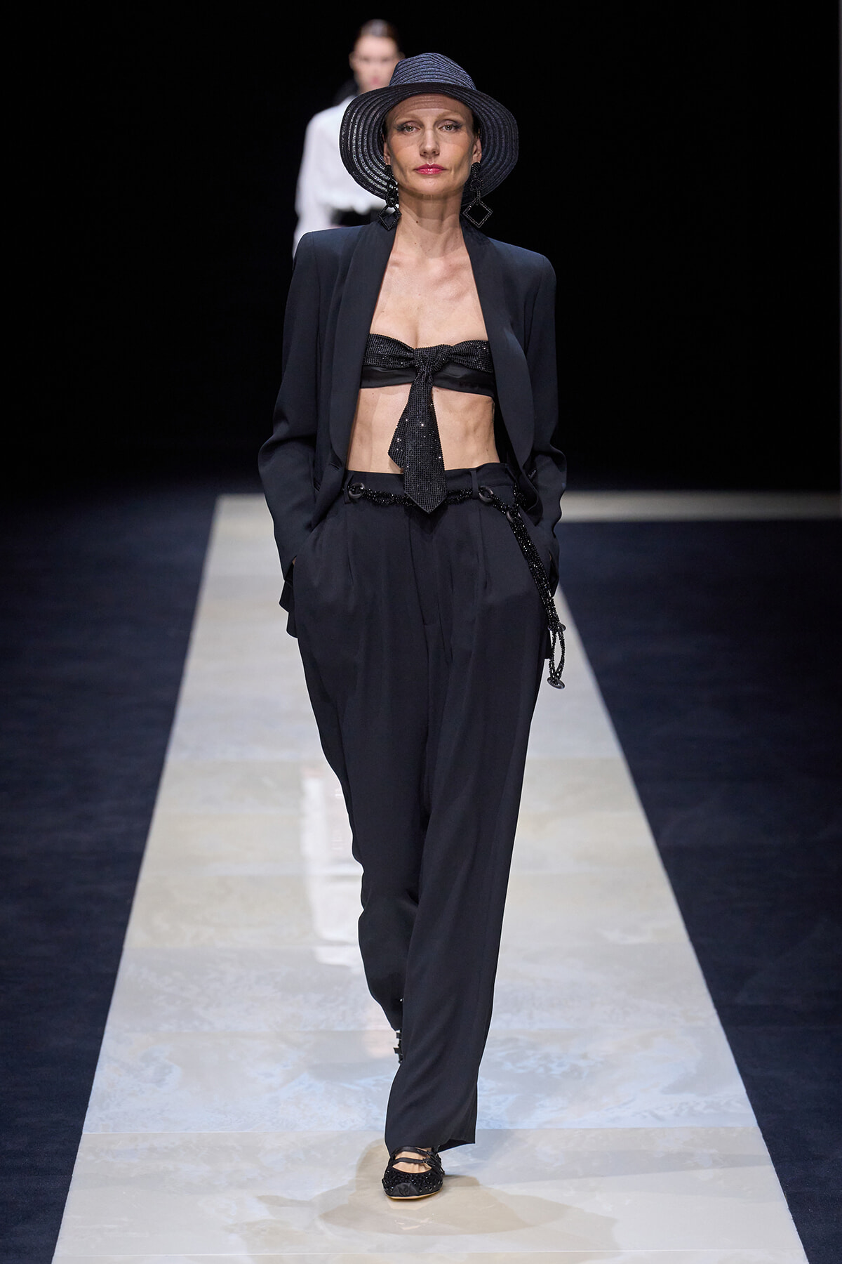 Model walks the runway in a black suit with a bralette top, tie at the front, and a wide-brim hat with sculpted earrings.