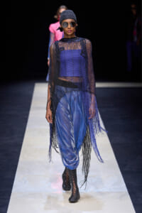 Model walking a runway in a blue metallic ensemble: sheer black mesh poncho with fringe over a blue top and pants, accessorized with a knit cap and oversized sunglasses.