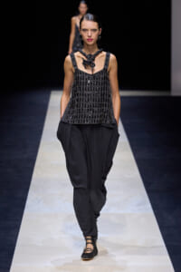 Model walks a fashion runway in an all-black look: beaded sleeveless top, loose black pants, chunky necklace, and slicked-back hair.