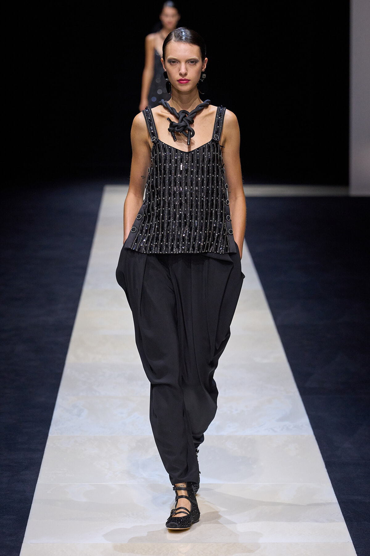 Model walks a fashion runway in an all-black look: beaded sleeveless top, loose black pants, chunky necklace, and slicked-back hair.