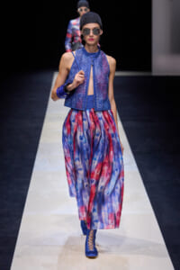 Model walks a fashion runway in a cobalt mesh sleeveless top with a keyhole front, tie-dye blue and red wide-leg pants, blue lace-up boots, and a dark knit cap with sunglasses.