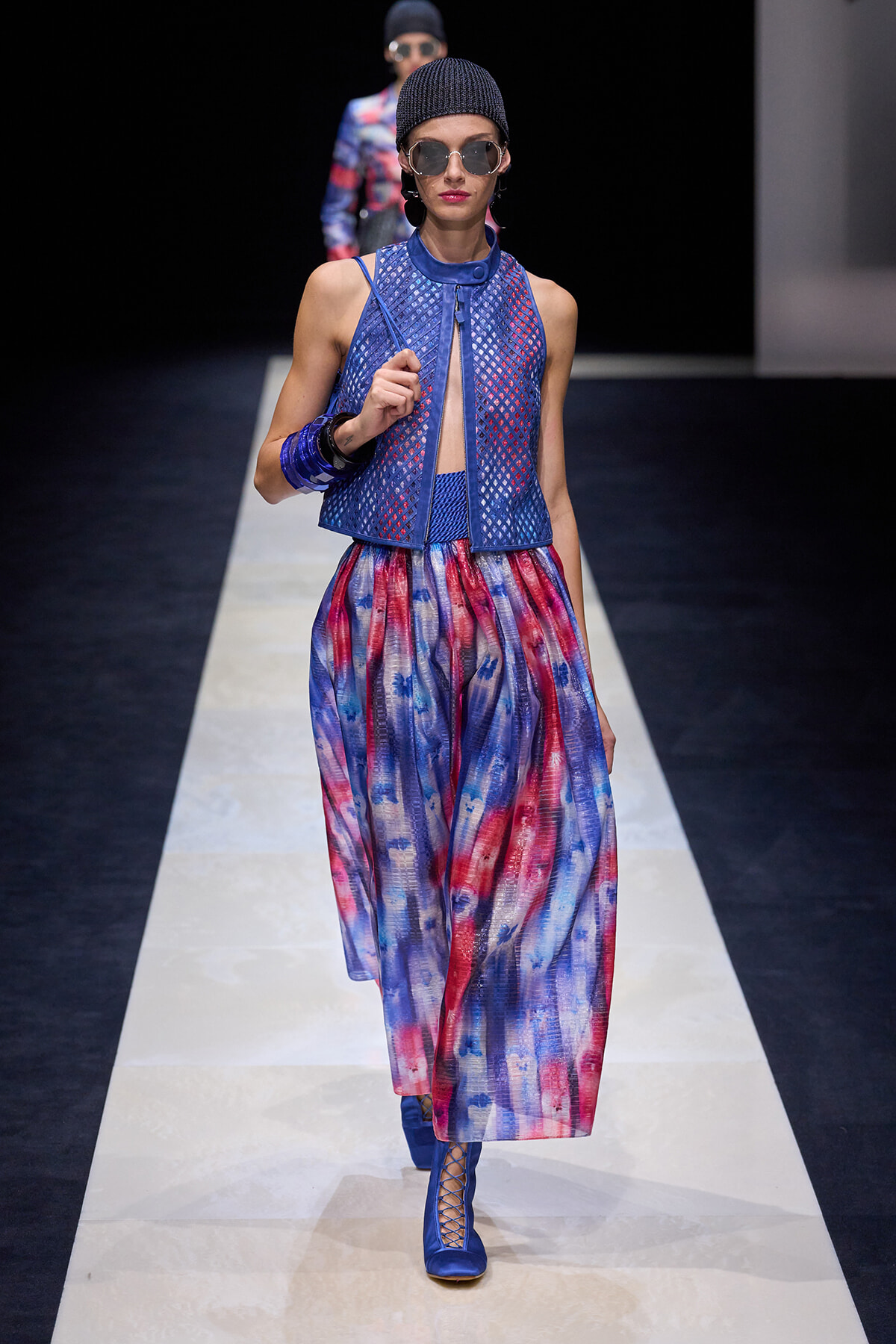 Model walks a fashion runway in a cobalt mesh sleeveless top with a keyhole front, tie-dye blue and red wide-leg pants, blue lace-up boots, and a dark knit cap with sunglasses.