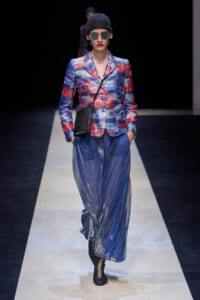 Female model on a runway wearing a colorful plaid blazer, blue metallic wide‑leg pants, black knit beanie, oversized sunglasses, and a small black crossbody bag.