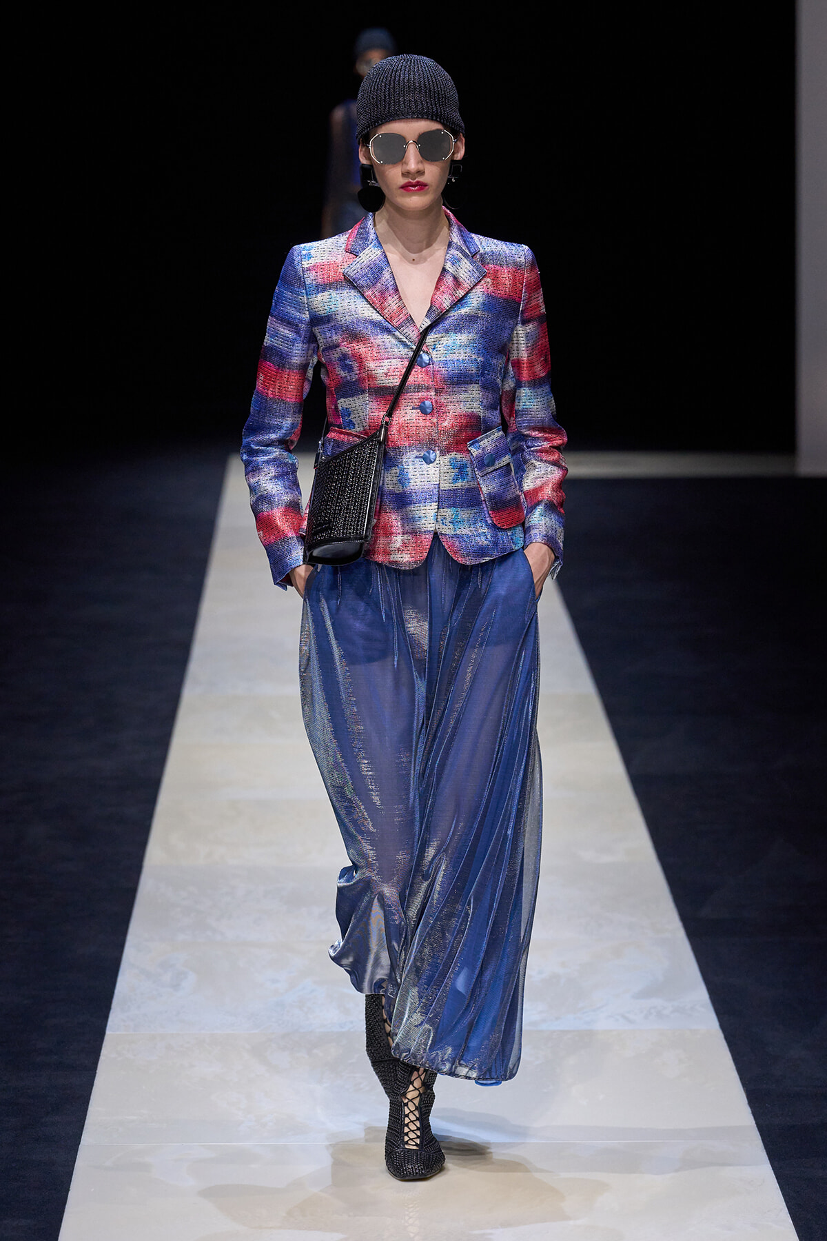 Female model on a runway wearing a colorful plaid blazer, blue metallic wide‑leg pants, black knit beanie, oversized sunglasses, and a small black crossbody bag.