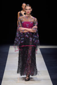Model on runway wearing a sheer black cape with blue florals over a magenta bodice and a beaded black skirt.