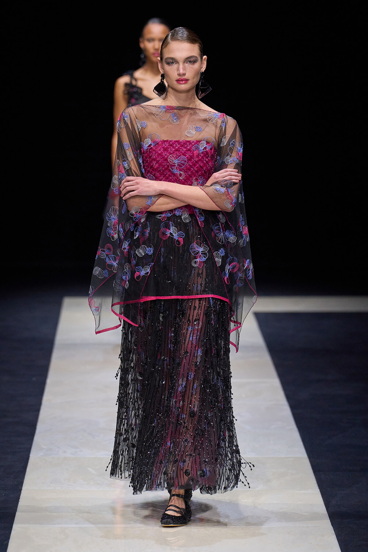 Model on runway wearing a sheer black cape with blue florals over a magenta bodice and a beaded black skirt.