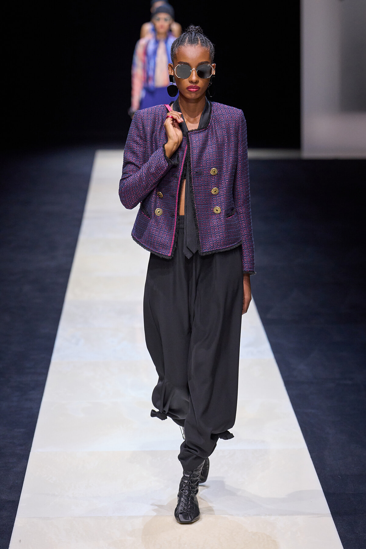 Model walking the runway in a purple-textured blazer with gold buttons, black draped pants, and round sunglasses.