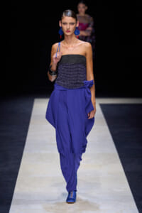 Model walks the runway in a strapless black top with cobalt blue draped pants, bow at the hip, matching blue earrings and shoes, dark backdrop.