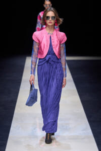 Model on a runway wearing a bright pink cropped jacket over a blue pleated dress, sunglasses, and pink earrings, carrying a blue handbag.