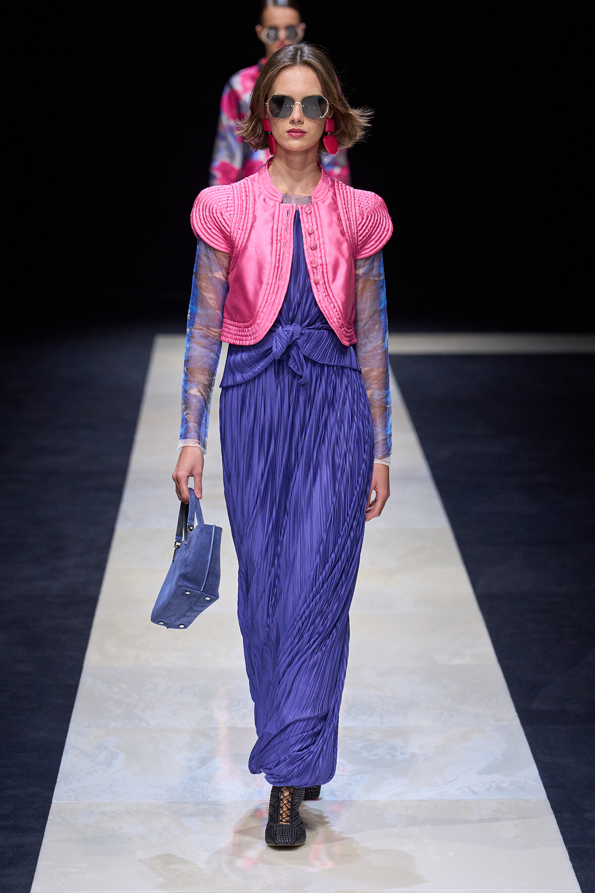 Model on a runway wearing a bright pink cropped jacket over a blue pleated dress, sunglasses, and pink earrings, carrying a blue handbag.