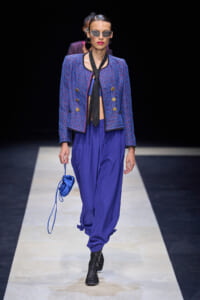 Model on a runway wearing a cobalt blue tweed cropped jacket with gold buttons, matching loose blue pants, a black necktie, black lace-up boots, sunglasses, and a small blue handbag.
