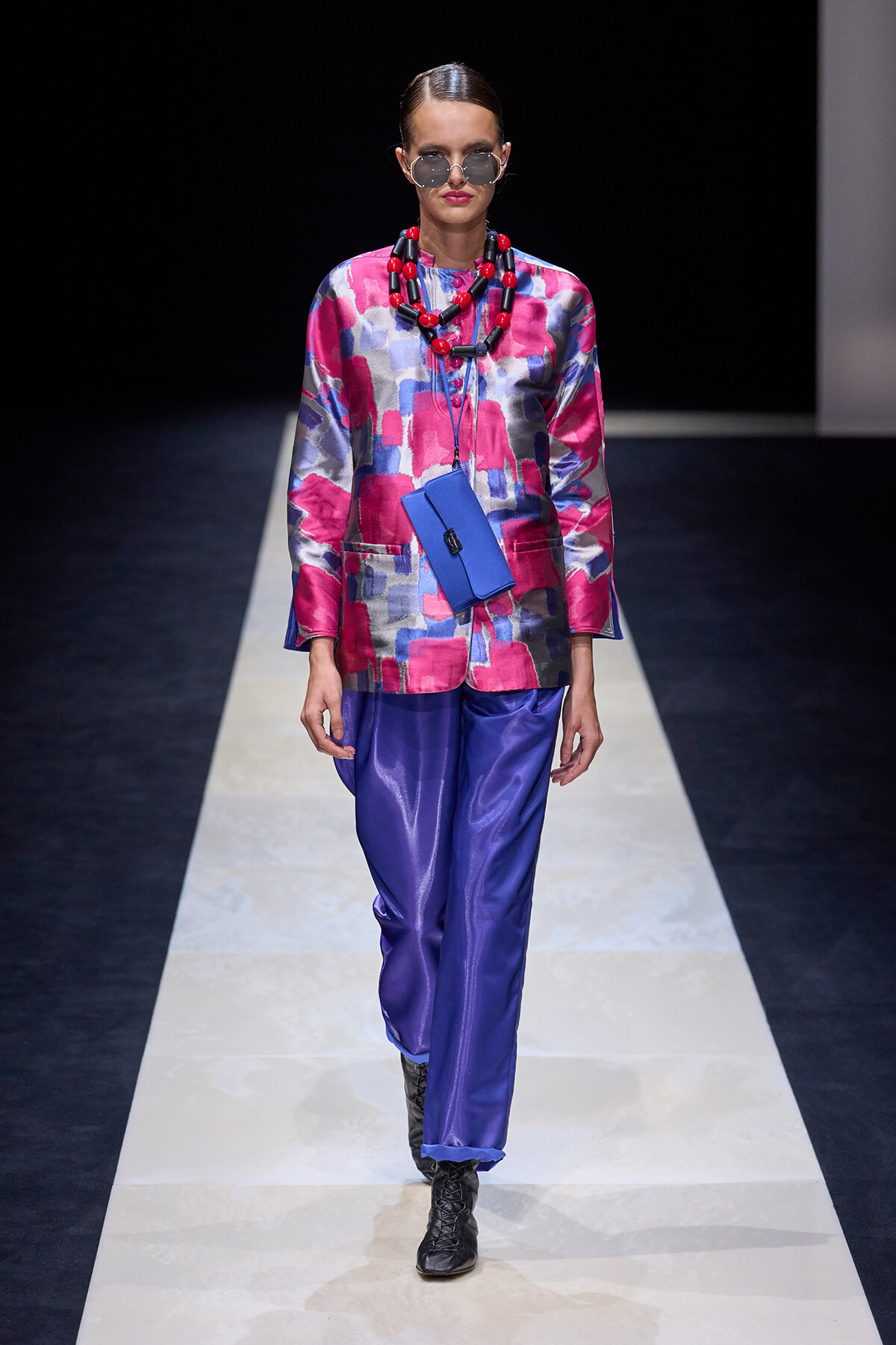 Model on a runway wearing a pink‑purple abstract blouse, blue satin pants, chunky red necklace, blue crossbody pouch, and round sunglasses.