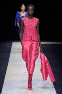 Model wearing a bright pink beaded sleeveless top and satin wide-leg pants, holding a matching jacket as she walks a runway.