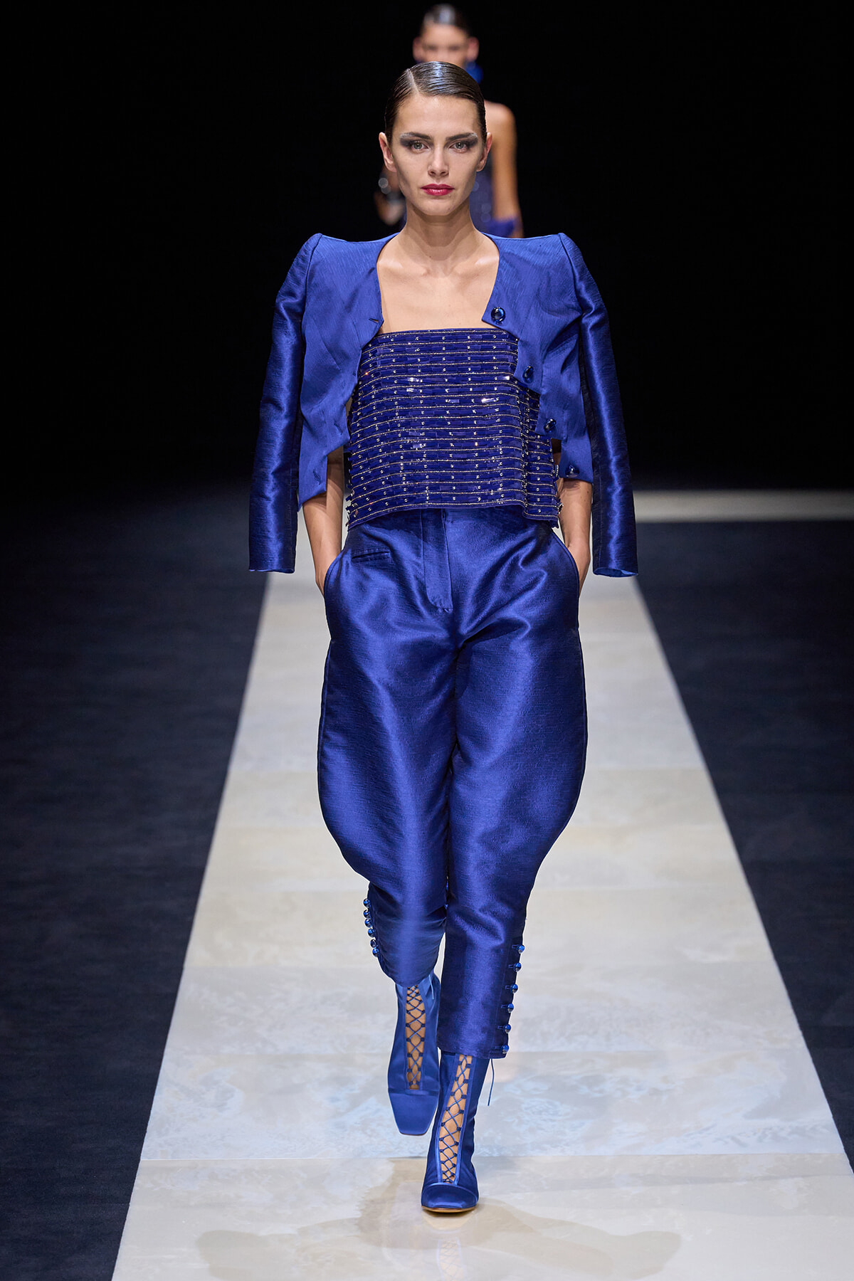 Model in a cobalt blue satin pantsuit with cropped beaded corset top, walking a runway on a light-colored catwalk.