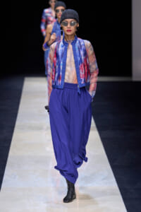 Model walks a fashion runway in a sheer multicolor blouse, cobalt jacket, and matching wide blue pants with black ankle boots and sunglasses.