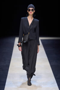 Model walking a runway in a dark tailored blazer, loose black pants, knit beanie, oversized sunglasses, and a small crossbody bag with a glittery finish.