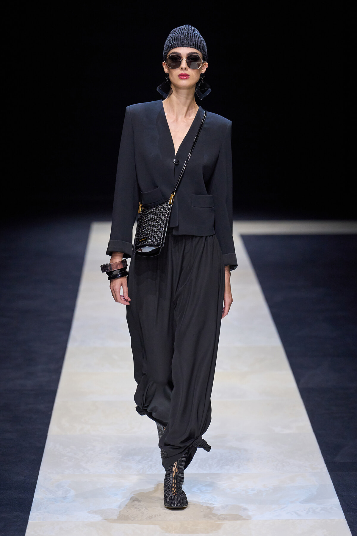 Model walking a runway in a dark tailored blazer, loose black pants, knit beanie, oversized sunglasses, and a small crossbody bag with a glittery finish.