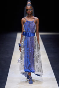 Model walks a runway wearing a blue and silver floral midi dress with a wide cobalt belt and blue sandals, accessorized with sunglasses and bracelets.