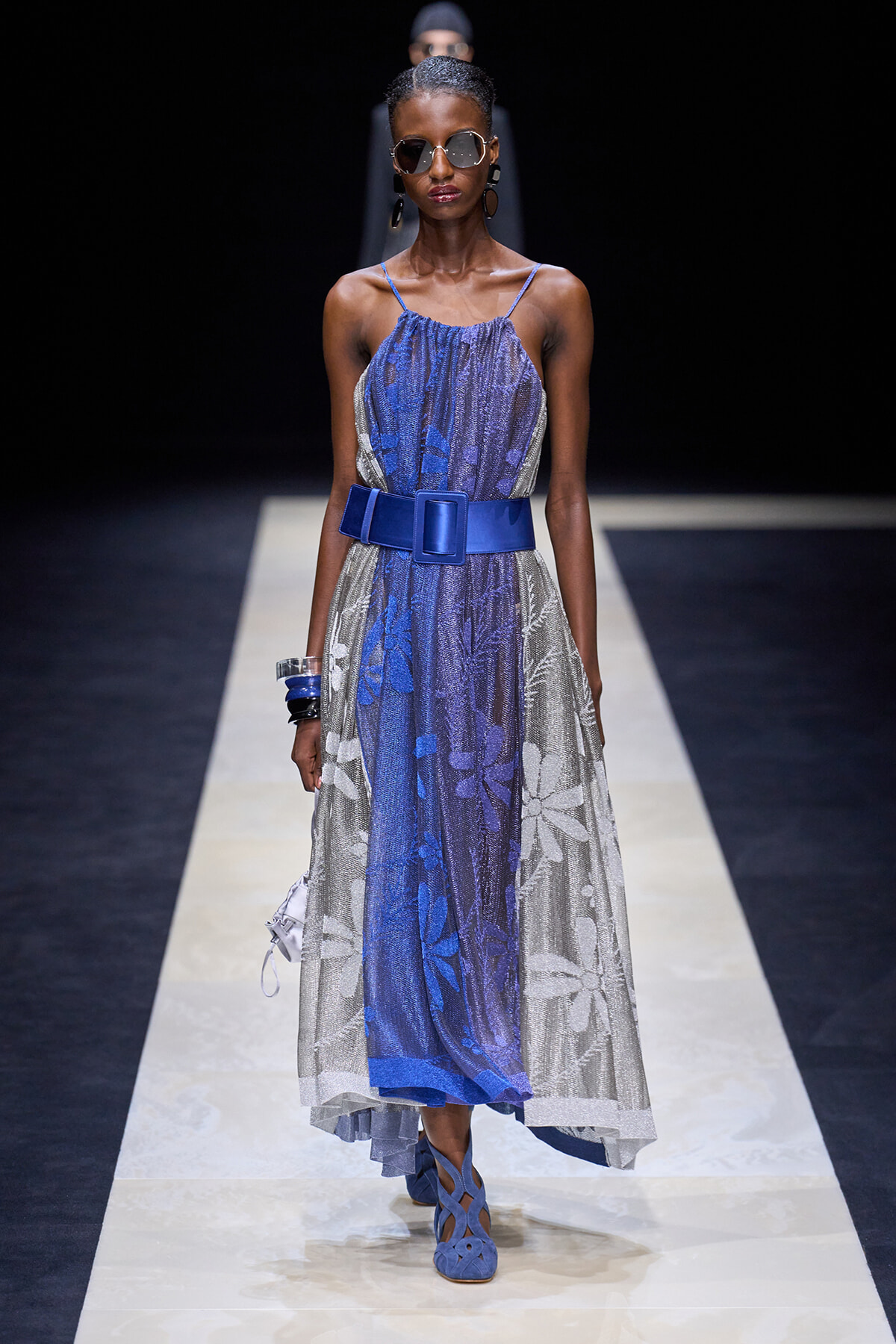 Model walks a runway wearing a blue and silver floral midi dress with a wide cobalt belt and blue sandals, accessorized with sunglasses and bracelets.