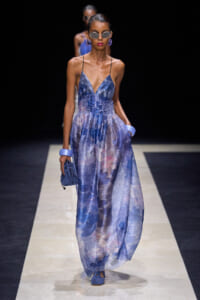 Model walking on a runway in a blue patterned maxi dress with thin straps and oversized sunglasses, carrying a blue clutch and wearing purple earrings and bracelets.