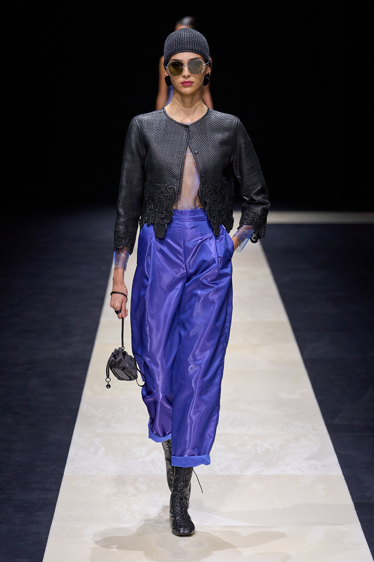 Runway model wearing a textured black jacket with a sheer center, paired with electric purple pants, black boots, a knit beanie, sunglasses, and a small bag on the runway.