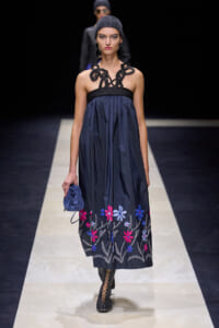 Model walking runway in navy strapless dress with black braided neckline and colorful floral embroidery at hem, paired with a blue clutch and black lattice boots.