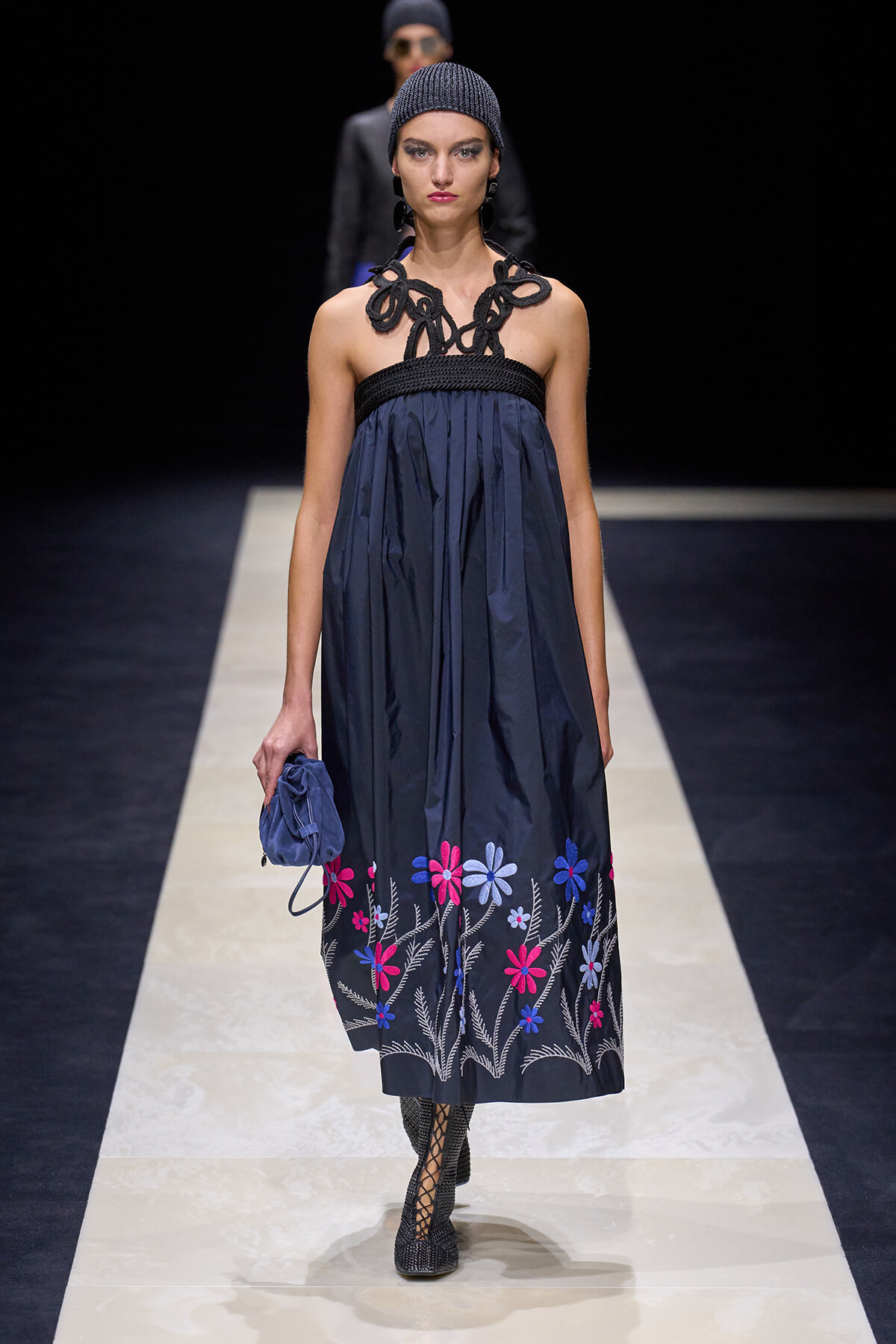Model walking runway in navy strapless dress with black braided neckline and colorful floral embroidery at hem, paired with a blue clutch and black lattice boots.