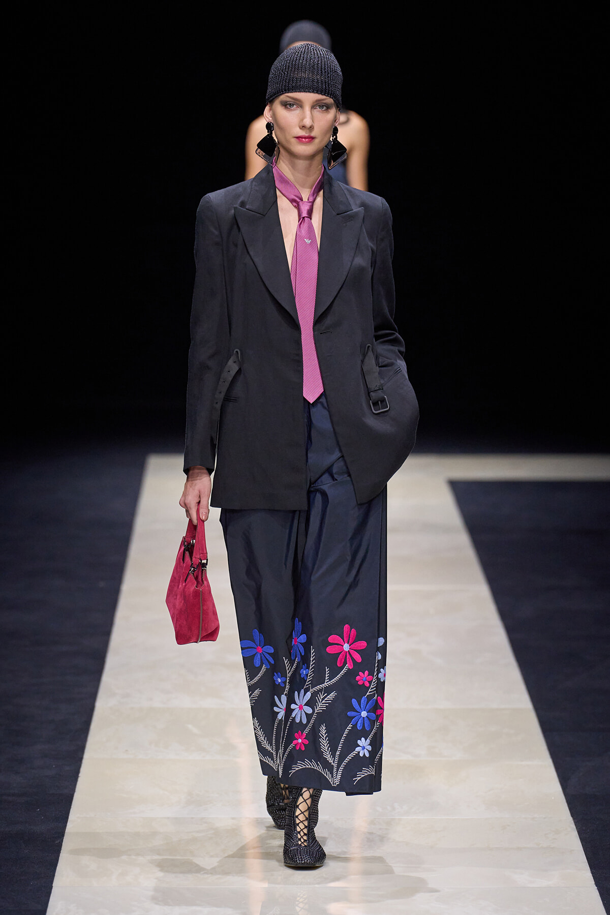 Model walking a fashion runway in a dark blazer, pink tie, navy floral-embroidered skirt, knit cap, and pink handbag.