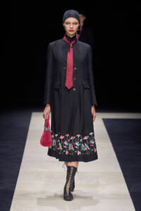 Model on a runway wearing a long black coat with colorful floral embroidery at the hem, a red tie, a dark beanie, and a small red handbag.