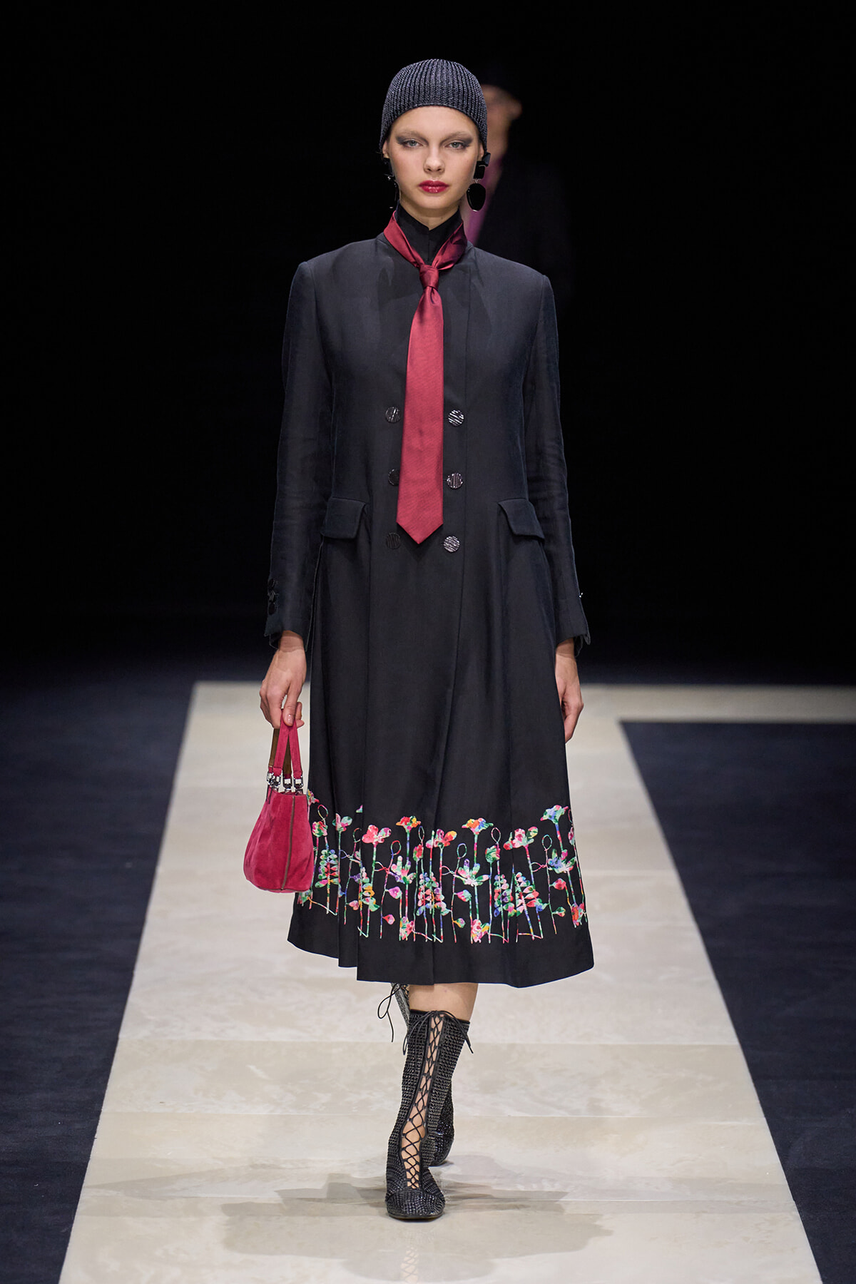 Model on a runway wearing a long black coat with colorful floral embroidery at the hem, a red tie, a dark beanie, and a small red handbag.