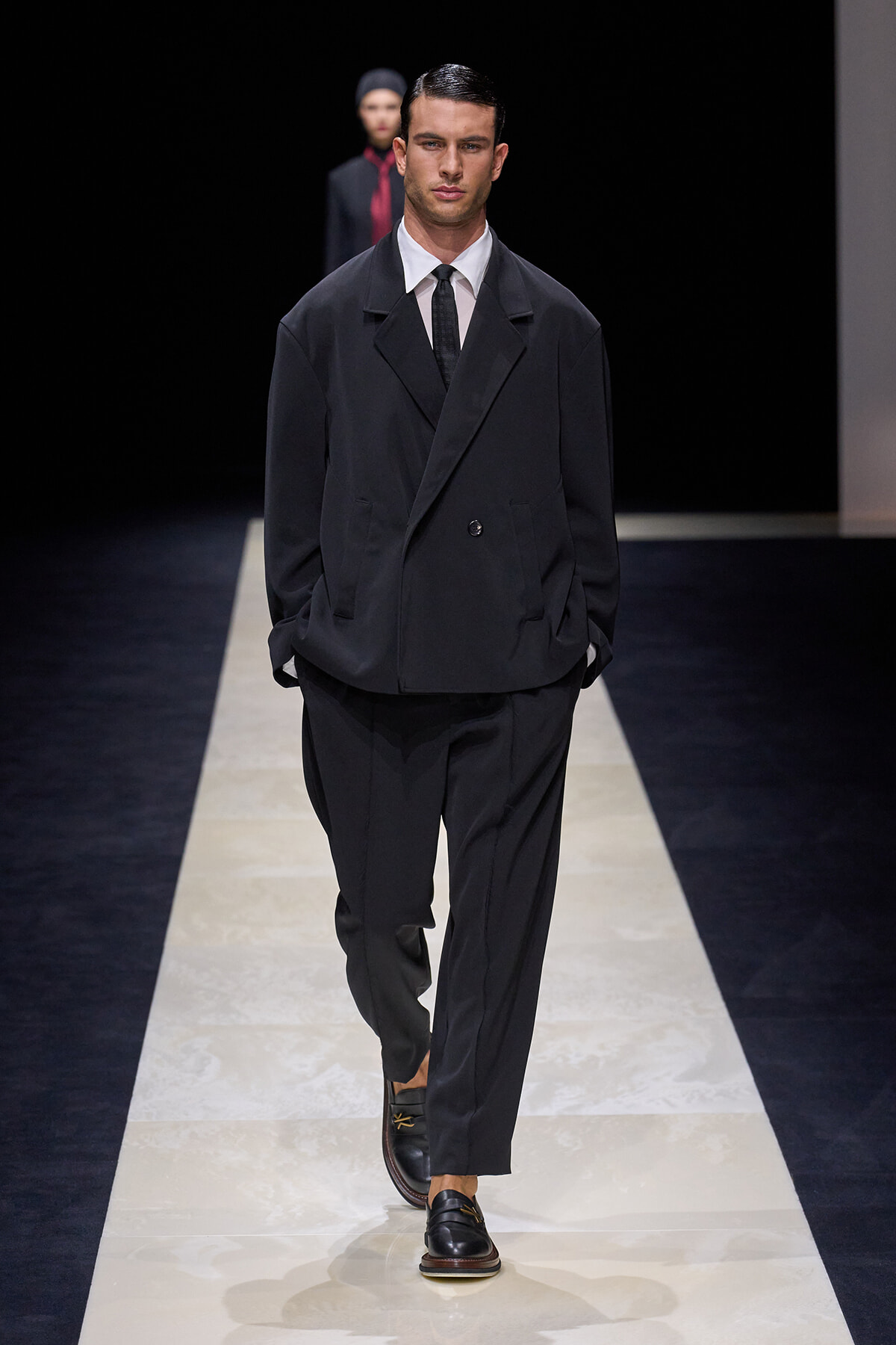 Male model walks a minimalist runway in an oversized black suit jacket, matching trousers, white shirt and black tie, hands in pockets.