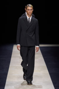 Male model wearing a dark double-breasted suit with a white shirt and black tie, walking down a fashion runway.