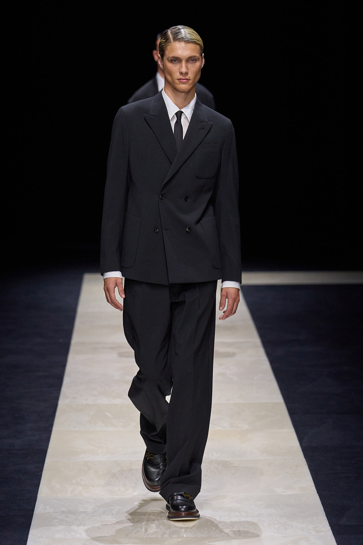 Male model wearing a dark double-breasted suit with a white shirt and black tie, walking down a fashion runway.