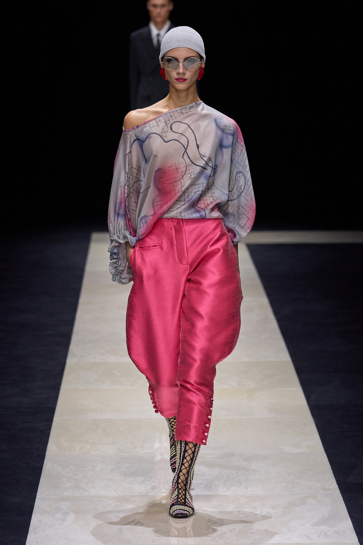 Runway model wearing an off-shoulder gray abstract-print blouse with loose sleeves and bright pink satin pants, paired with patterned lace-up boots and a white knit cap.